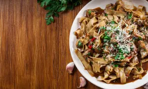 Pyszne makaron tagliatelle z grzybami i parmezanem, ozdobione natką pietruszki. Obok leżą ząbki czosnku.