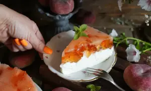 Pyszny sernik z jogurtów greckich z brzoskwiniową galaretką, ozdobiony miętą. Obok świeże brzoskwinie i kwiaty.