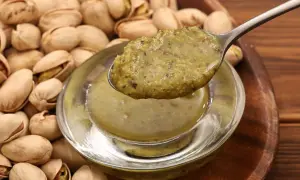 Łyżka z gęstą, zieloną masą pistacjową nad miseczką, w tle stos pistacji.
