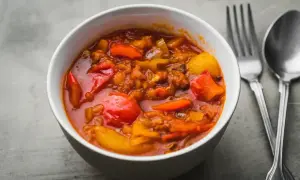 Aromaticzne leczo warzywne w białej misce, z widocznymi kawałkami papryki, pomidorów i ziemniaków. Obok sztućce.