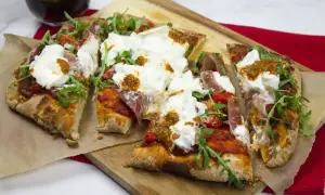 Pyszna pinsa z burratą, rukolą, pomidorkami i szynką parmeńską, podana na desce.