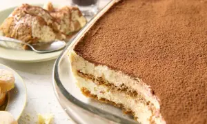 Pyszne tiramisu bez jajek, obficie posypane kakao, w szklanej formie. Na talerzyku porcja deseru i łyżeczka.