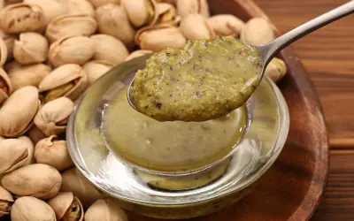 Łyżka z gęstą, zieloną masą pistacjową nad miseczką, w tle stos pistacji.