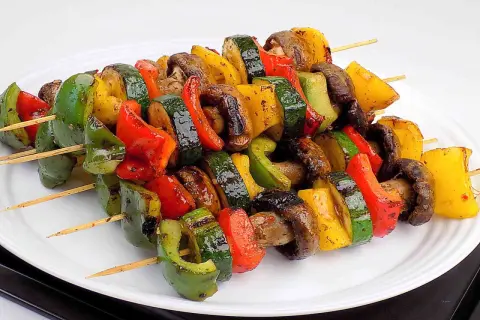 Kolorowe szaszłyki warzywne z papryką, cukinią i pieczarkami, grillowane do perfekcji, prezentują się apetycznie na białym talerzu.