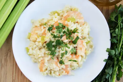 Kremowe risotto z krewetkami, posypane świeżą natką pietruszki, obok łodygi selera i oliwy.