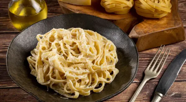 Pyszne fettuccine alfredo na czarnym talerzu, obok surowy makaron, ser i oliwa.