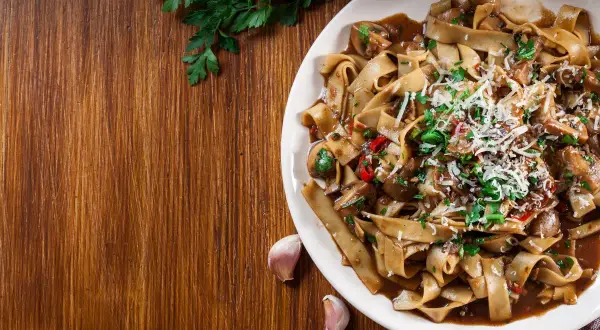 Pyszne makaron tagliatelle z grzybami i parmezanem, ozdobione natką pietruszki. Obok leżą ząbki czosnku.