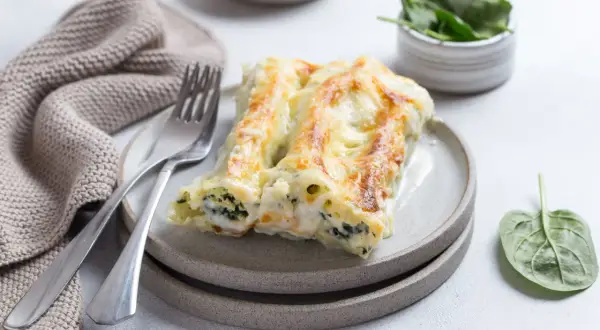 Pyszne cannelloni ze szpinakiem, zapieczone pod beszamelem i serem, na talerzu z widelcem i serwetką.