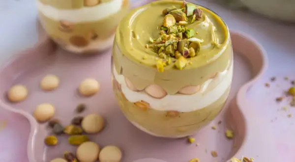 Pyszne tiramisu pistacjowe w szklance, przełożone kremem i biszkoptami, posypane orzechami pistacjowymi.