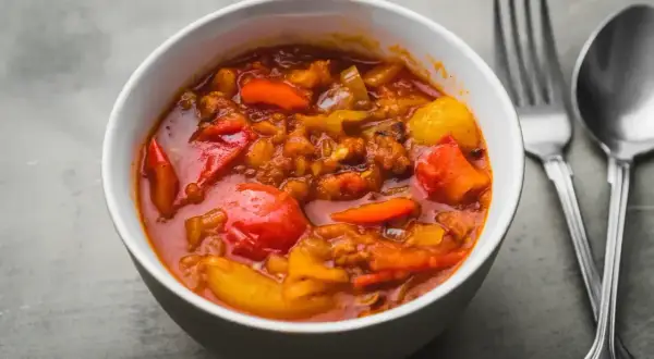 Aromaticzne leczo warzywne w białej misce, z widocznymi kawałkami papryki, pomidorów i ziemniaków. Obok sztućce.