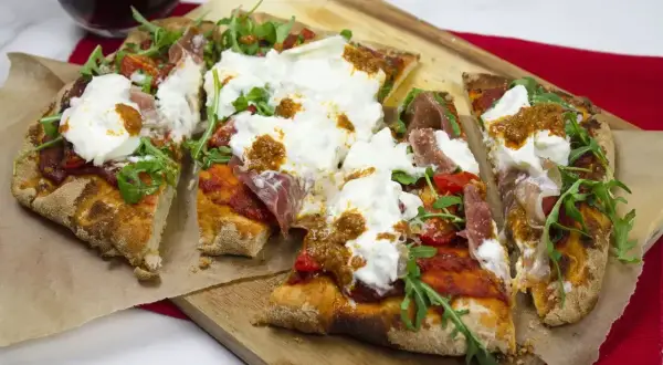 Pyszna pinsa z burratą, rukolą, pomidorkami i szynką parmeńską, podana na desce.