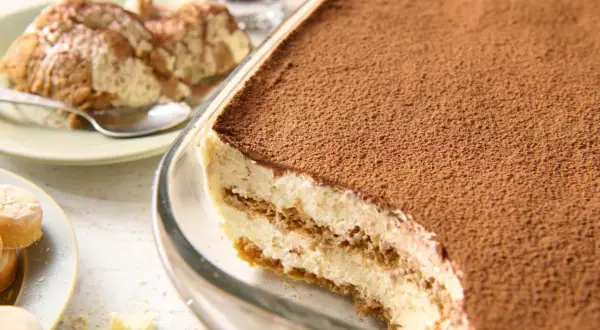 Pyszne tiramisu bez jajek, obficie posypane kakao, w szklanej formie. Na talerzyku porcja deseru i łyżeczka.