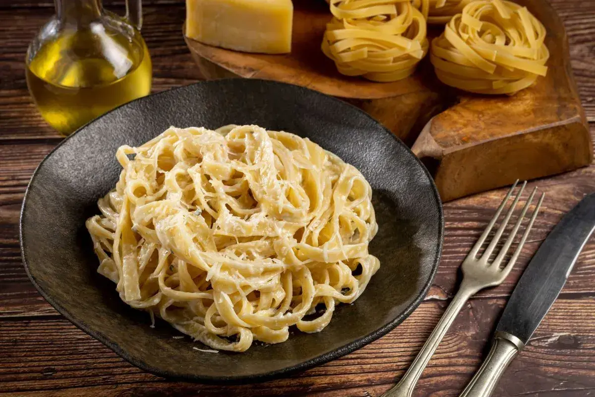 Pyszne fettuccine alfredo na czarnym talerzu, obok surowy makaron, ser i oliwa.