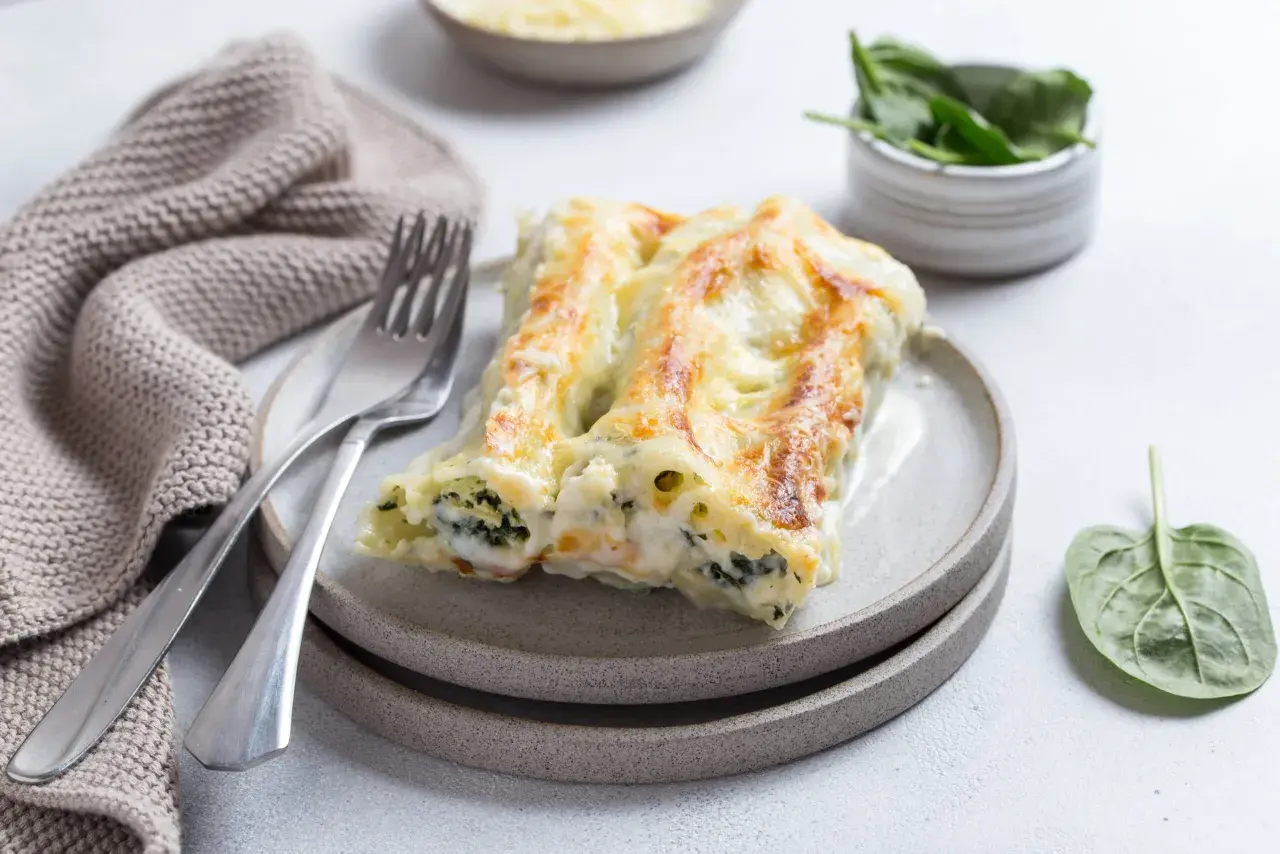 Pyszne cannelloni ze szpinakiem, zapieczone pod beszamelem i serem, na talerzu z widelcem i serwetką.
