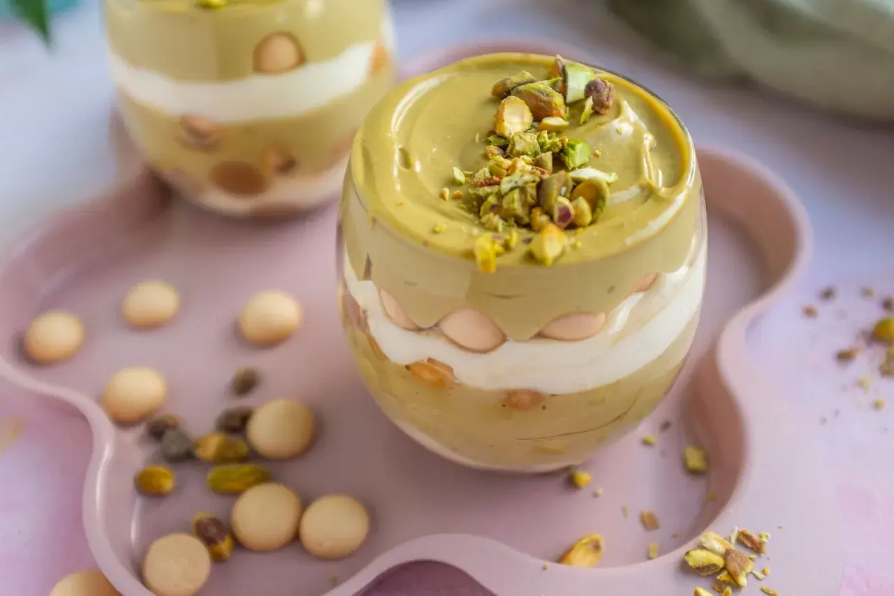 Pyszne tiramisu pistacjowe w szklance, przełożone kremem i biszkoptami, posypane orzechami pistacjowymi.