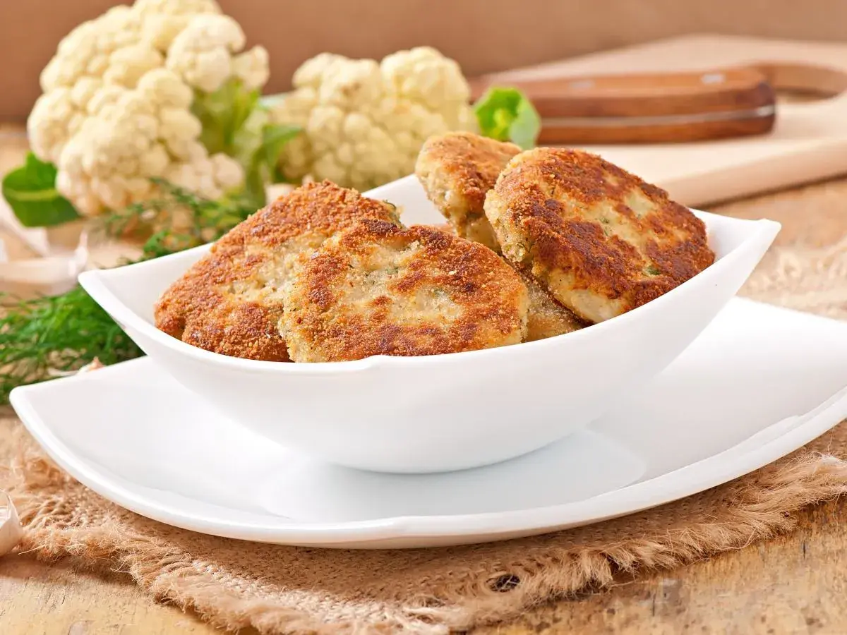 Złociste kotlety z kalafiora w białej misce, obok świeży kalafior i koperek. Pyszne danie wegetariańskie.