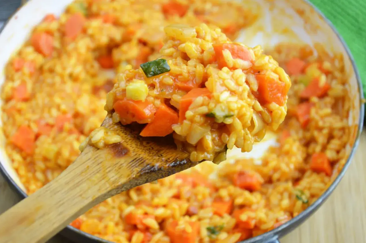 Kremowe risotto pomidorowe z kawałkami marchewki i cukinii, nabierane drewnianą łyżką.