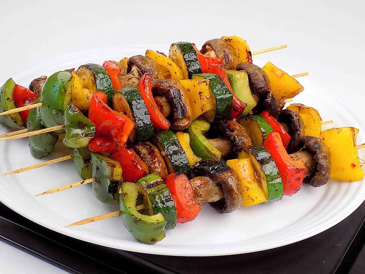 Kolorowe szaszłyki warzywne z papryką, cukinią i pieczarkami, grillowane do perfekcji, prezentują się apetycznie na białym talerzu.