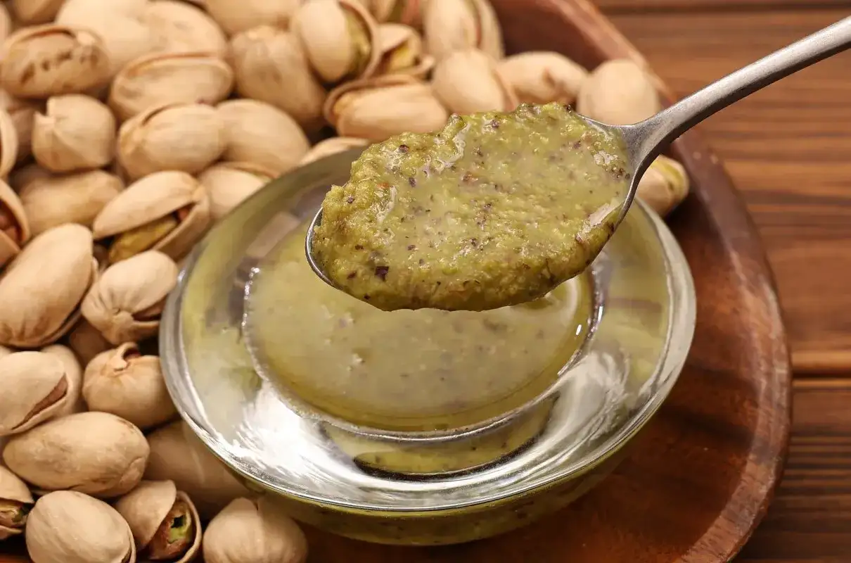 Łyżka z gęstą, zieloną masą pistacjową nad miseczką, w tle stos pistacji.