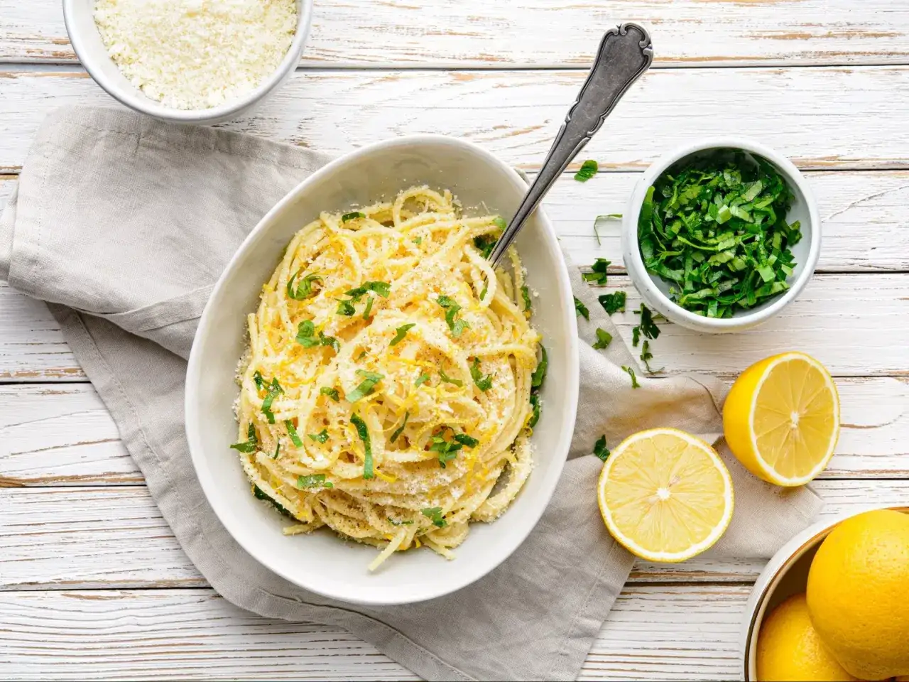 Aksamitne spaghetti al limone, posypane parmezanem i świeżą natką, z cytrynami w tle.