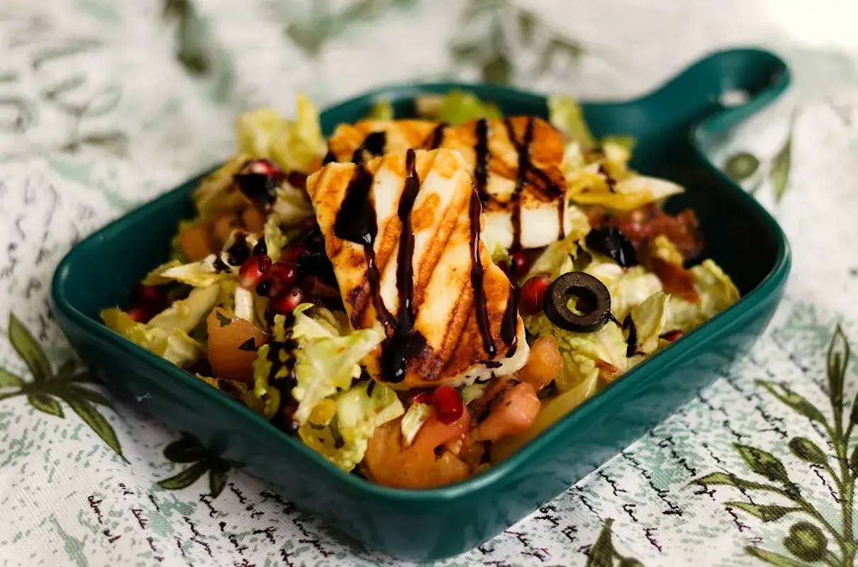 Sałatka z chrupiącą sałatą, pomidorami, oliwkami i granatem, zwieńczona grillowanym halloumi z piekarnika, polana sosem balsamicznym.