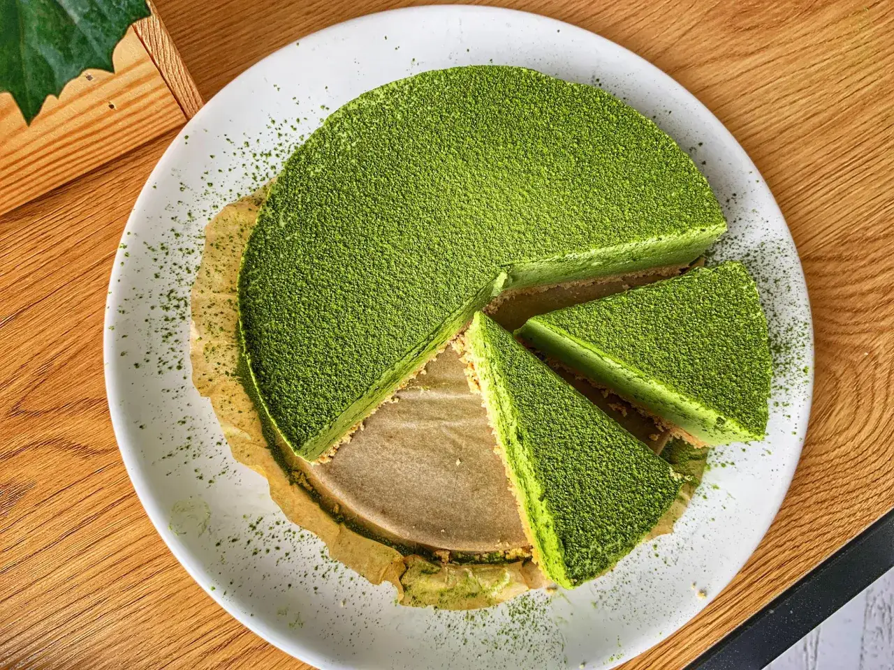 Zielony, wytrawny sernik matcha, posypany pudrem matcha, pokrojony na kawałki, na białym talerzu.