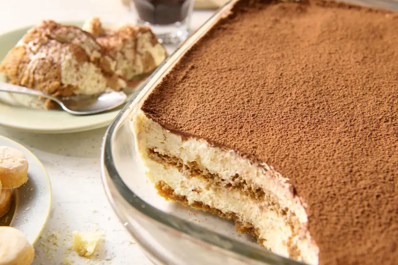 Pyszne tiramisu bez jajek, obficie posypane kakao, w szklanej formie. Na talerzyku porcja deseru i łyżeczka.