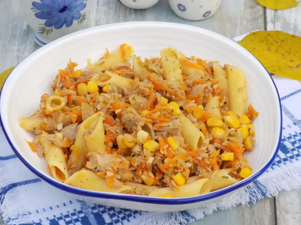 Penne z tuńczykiem, kukurydzą i startą marchewką w białej misce na niebiesko-białej serwetce.