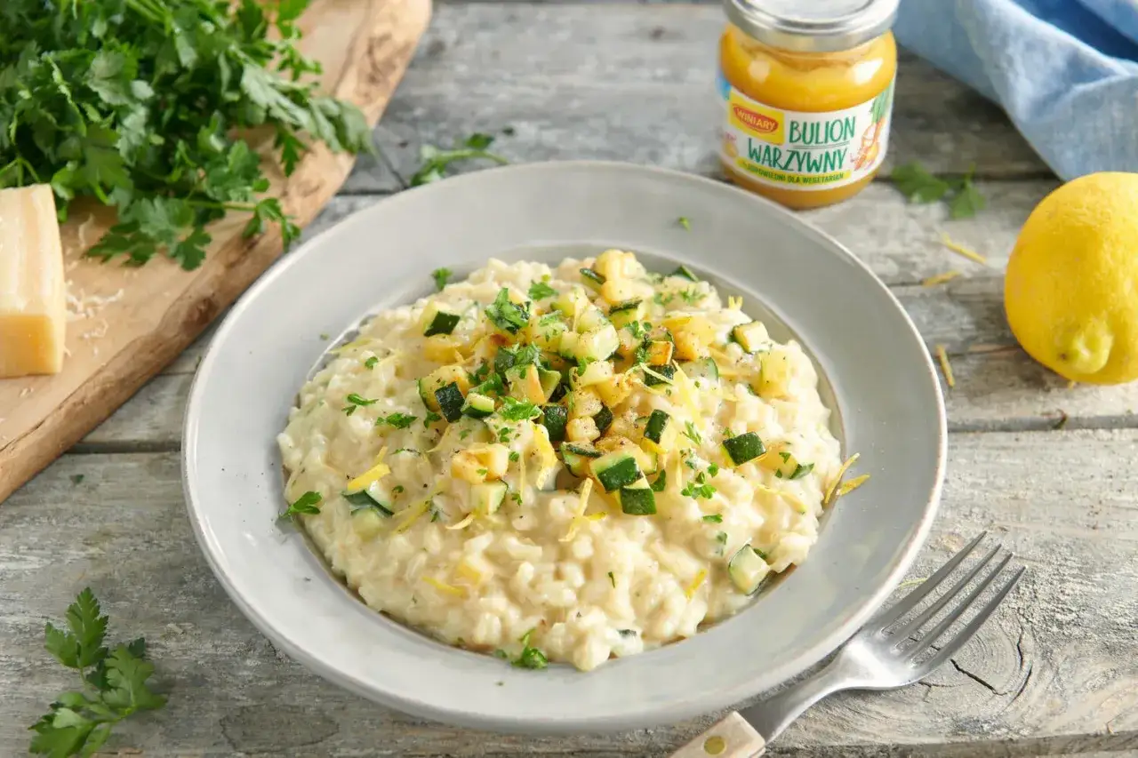 Kremowe risotto z cukinią, posypane świeżą natką pietruszki i skórką cytrynową. Obok kawałek parmezanu i cytryna.