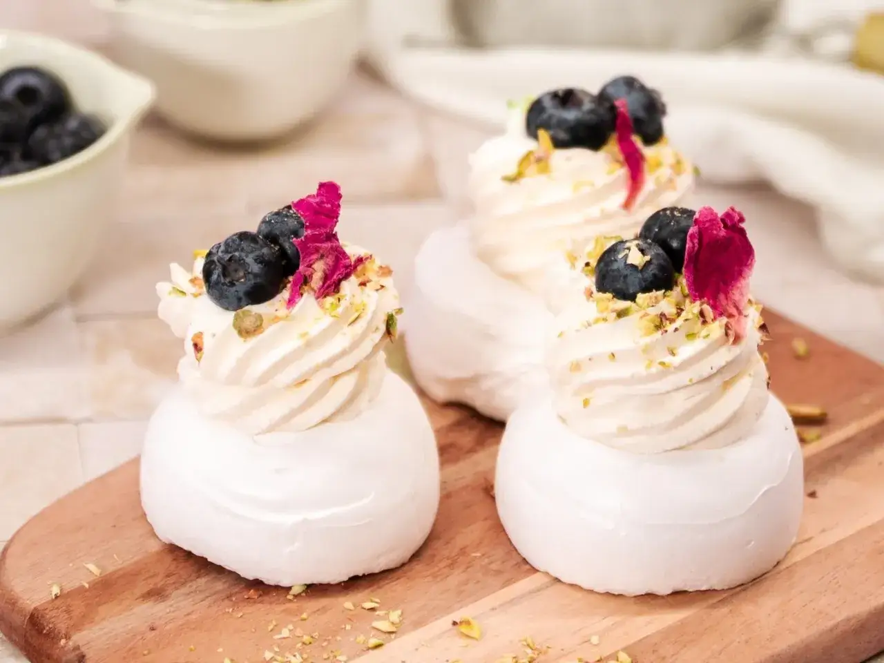 Trzy urocze mini pavlova z bitą śmietaną, borówkami, płatkami róży i pistacjami na drewnianej desce.