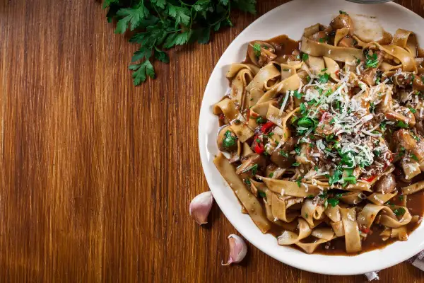 Pyszne makaron tagliatelle z grzybami i parmezanem, ozdobione natką pietruszki. Obok leżą ząbki czosnku.
