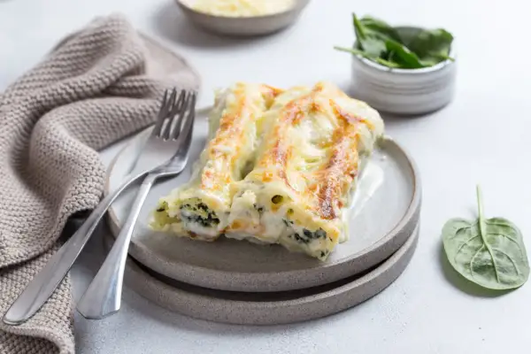 Pyszne cannelloni ze szpinakiem, zapieczone pod beszamelem i serem, na talerzu z widelcem i serwetką.