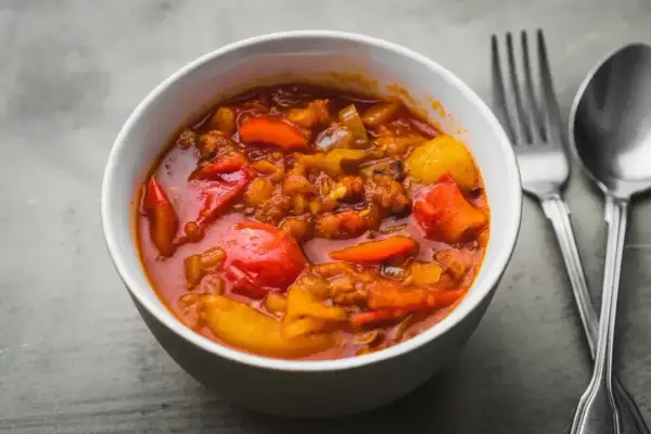 Aromaticzne leczo warzywne w białej misce, z widocznymi kawałkami papryki, pomidorów i ziemniaków. Obok sztućce.
